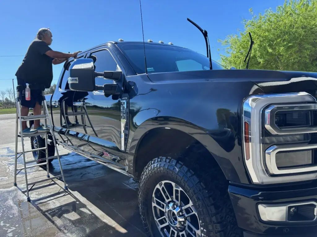 The owner of Adame and Son's is on a ladder cleaning the top of a black Ford dually truck